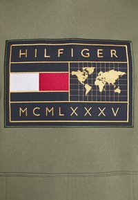 Felpa verde oliva con un patch blu navy che presenta il logo "HILFIGER", una mappa del mondo in oro e strisce orizzontali rosse, bianche e blu.