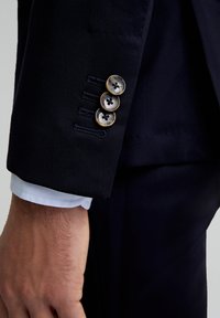 Manga de chaqueta de traje azul marino con tres botones marrones de cuatro orificios, ojales cosidos y un puño de camisa azul claro debajo.