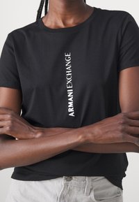 Czarna koszulka z krótkim rękawem wykonana z bawełny, z pionowym białym napisem "ARMANI EXCHANGE" umieszczonym na środku przodu.