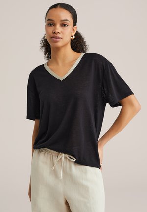 Femeie cu păr creț, purtând cercei aurii tip cerc, o bluză neagră cu decolteu în V și margine metalică, și pantaloni bej cu șnur, stând cu mâna pe șold.