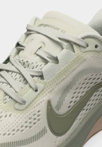 Chaussure de course Nike Pegasus 42 beige clair avec dessus en mesh, lacets détaillés et logo swoosh vert olive sur le côté.