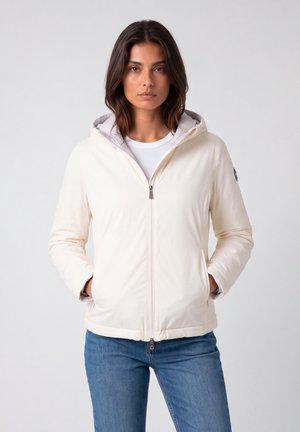 Donna con capelli scuri che indossa una giacca bianca con cappuccio e zip e jeans blu, in piedi con le mani in tasca su uno sfondo semplice.
