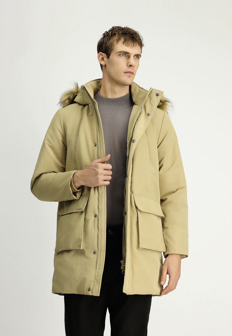 Hombre vistiendo una parka beige con capucha y adornos de piel, suéter gris y pantalones negros, de pie contra un fondo claro y sencillo, mirando de lado.