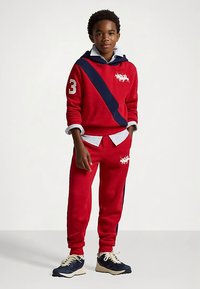 Rapaz de pé com um fato de treino vermelho com riscas azul-escuro, camisa branca por baixo e ténis azul-escuro com atacadores brancos, contra um fundo simples.