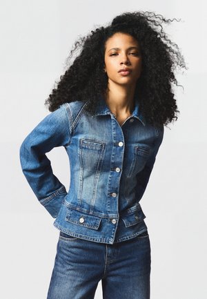 Femme aux cheveux bouclés portant une veste en jean bleue et un jean, debout, les mains derrière le dos, devant un fond clair uni.