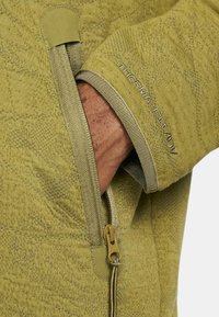 Jachetă verde măslinie dintr-un material texturat, cu un buzunar cu fermoar. Prezintă manșete elastice și înscrisul "THERMA-FIT ADV" brodat pe mânecă.