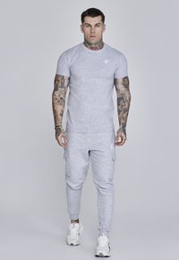 Svetlo siva športna majica z okroglo ovratnico in logotipom, v kombinaciji s sorodnimi sivimi cargo hlačami s stranskimi žepi in rebrastimi manšetami.