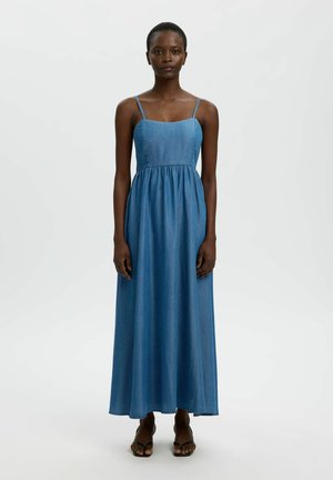 Kobieta stoi zwrócona przodem, ubrana w niebieską denimową suknię maxi bez rękawów z cienkimi ramiączkami i czarne, otwarte na palce buty na obcasie na jasnym, jednolitym tle.