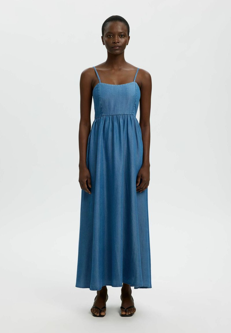 Femme debout, faisant face, portant une robe maxi en jean bleu sans manches avec des fines bretelles et des talons ouverts noirs sur un fond clair uni.
