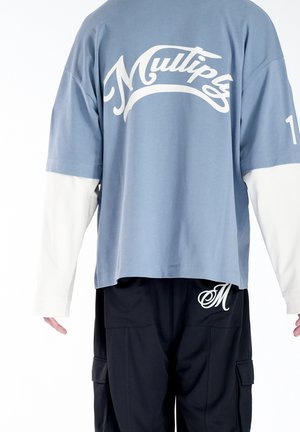 Niebieski, oversize'owy T-shirt z krótkim rękawem, pod spodem białe długie rękawy, na piersi napis „Multiply”, w połączeniu z czarnymi spodniami z logo „M”.