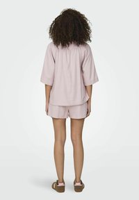 Lichtroze, kortmouwtop met een losse pasvorm en een plooi aan de achterkant, gecombineerd met bijpassende shorts. Gekenmerkt door casual bruine sandalen met roze accenten.