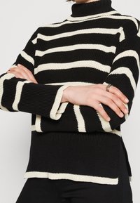 Pull tricoté à rayures noires et blanches avec un col montant, une texture côtelée et des fentes latérales à l'ourlet, porté avec les bras croisés.