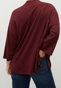 Burgunderfarbener, locker sitzender Oberteil mit 3/4-Ärmeln, einer hohen Halslinie und seitlichen Schlitzen, kombiniert mit dunkelblauen Jeans. Glattes, strukturiertes Material.