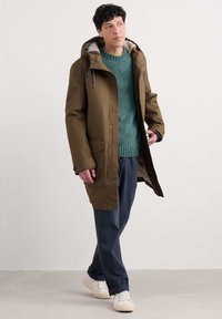 Vyras eina patalpoje, vilkintis alyvuogių spalvos parką su pūkine pamušalu, mėtų spalvos megztinį, tamsiai mėlynas kelnes ir baltus sportinius batelius prieš paprastą sienos foną.