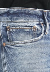 Ljusblå denimjeans med en framficka som har kopparfärgade nitar, synlig sömnad och en sliten textur vid midjan.