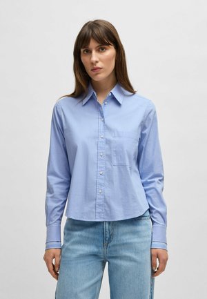 Camicia azzurro chiaro con maniche lunghe, colletto a punte, taschino sul petto e orlo leggermente accorciato. Tessuto dalla texture liscia.