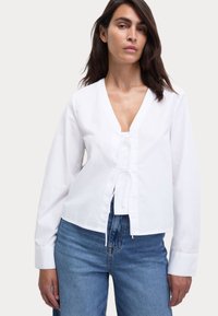 Blusa bianca a maniche lunghe con scollo a V e fiocco frontale. Realizzata in cotone, presenta una vita aderente ed è abbinata a jeans blu a vita alta.