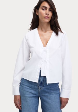 Blusa bianca a maniche lunghe con scollo a V e fiocco frontale. Realizzata in cotone, presenta una vita aderente ed è abbinata a jeans blu a vita alta.