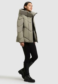 Gepolsterte olivgrüne Pufferjacke mit hohem Kragen, ausgestattet mit einem Zickzackmuster und einem Reißverschluss vorne. Kombiniert mit schwarzen Leggings und knielangen schwarzen Stiefeln.