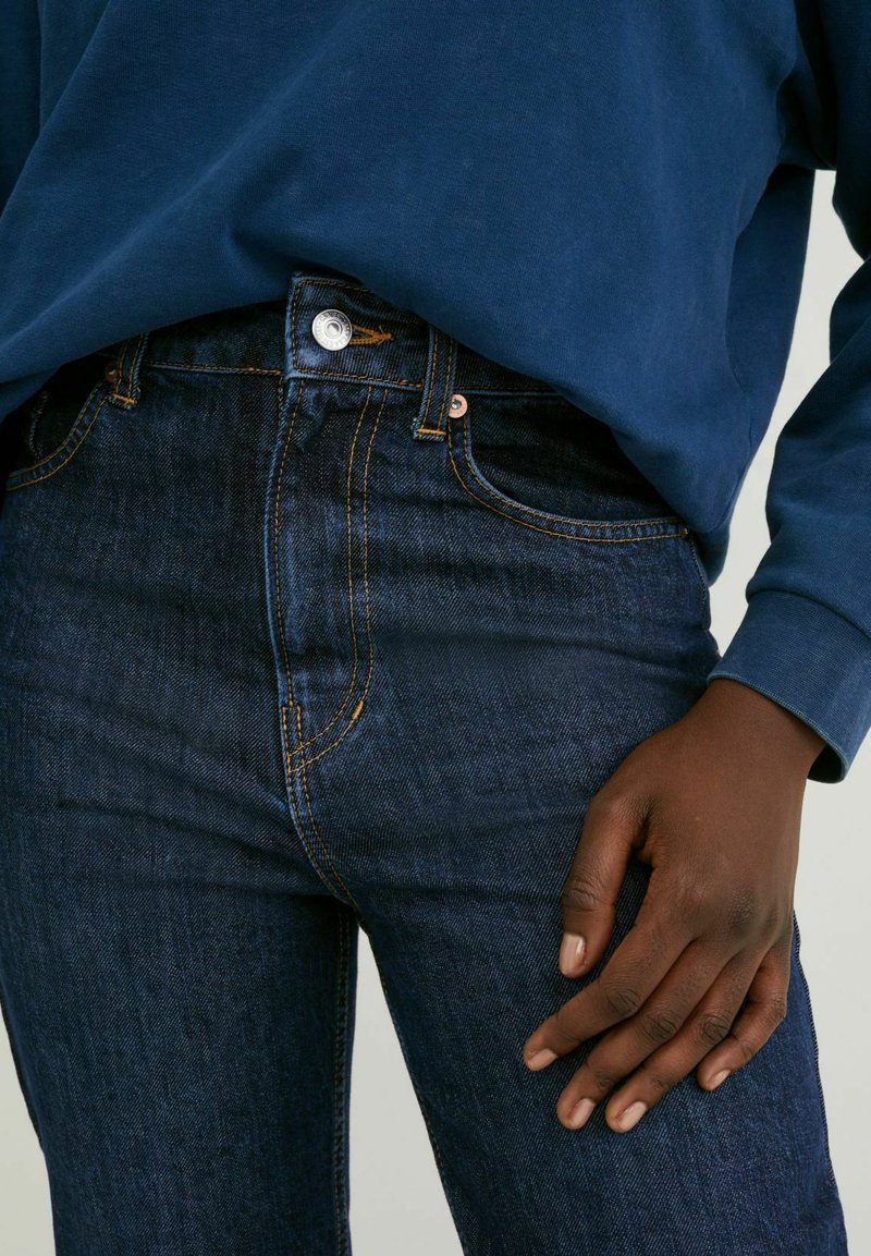 Jean en denim bleu foncé avec une taille haute, des poches avant et des coutures orange contrastantes. Associé à une chemise à manches longues bleu marine.