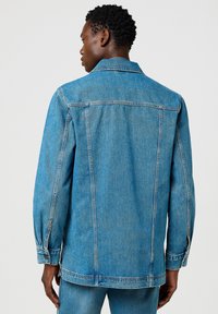 Chaqueta de denim en azul claro, con un cuello clásico, mangas largas, puños abotonados y detalles de costura visibles en la parte posterior.