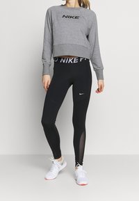 Donna che indossa un felpa grigia Nike, leggings neri Nike con pannelli in rete e scarpe da ginnastica bianche, in piedi contro uno sfondo neutro.