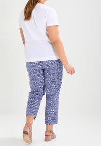 White short-sleeve top paired with blue patterned ankle-length trousers, featuring a geometric design and light fabric. Brown block-heeled shoes.
