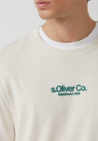 Cream-colored sweatshirt with ribbed crew neckline, featuring green embroidered text "s.Oliver Co. Established 1969" on the front.