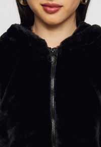 Svart imitationpälsjacka med dragkedja fram, med rund dragkedjetag och en mjuk, texturerad yta. Huva detaljer är synliga.