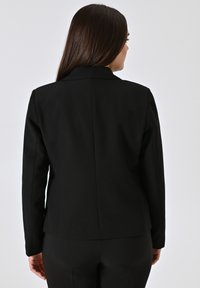 Schwarze, taillierte Blazerjacke mit schmaler Passform, elongated Silhouette und klaren Linien. Verfügt über einen Reverskragen und eine glatte Stoffqualität.