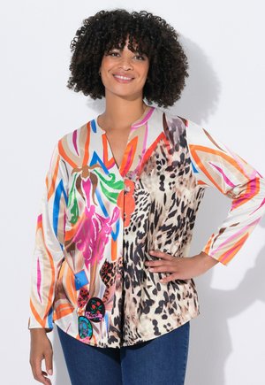 Femme aux cheveux bouclés sourit, portant un blouse à manches longues colorée avec des motifs floraux et animaliers, associée à un jean bleu.