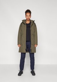 Oliven grøn parka jakke med hætte, frontlynlås og store lommer, parret med marineblå bukser og sorte sko. Glat tekstur og afslappet design.