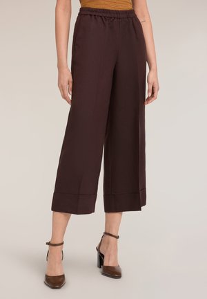 Kobieta w ciemnobrązowych, szerokich spodniach capri z wysokim stanem, połączonych z brązowymi butami na słupku z paskiem na kostkę, na neutralnym tle.