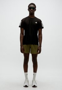 Μαύρο αθλητικό t-shirt με λευκές ρίγες, χακί σορτς, λευκές κάλτσες και γκρι παπούτσια. Ο μοντέλο φοράει σκούρα γυαλιά ηλίου, στέκεται μπροστά σε απλό φόντο.