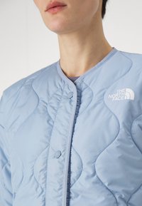 Ljusblå vadderad jacka med ribbad krage och tryckknappar, med den vita "The North Face"-logotypen på vänster sida.