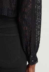 Blouse en dentelle noire à manches longues avec un motif texturé. Les poignets sont fermés par des boutons. Associée à un pantalon noir ajusté.