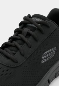 Svart atletisk sko med en teksturert overflate, runde snørebånd og en subtil logo. Designet inkluderer ventilasjonshull og en polstret såle.