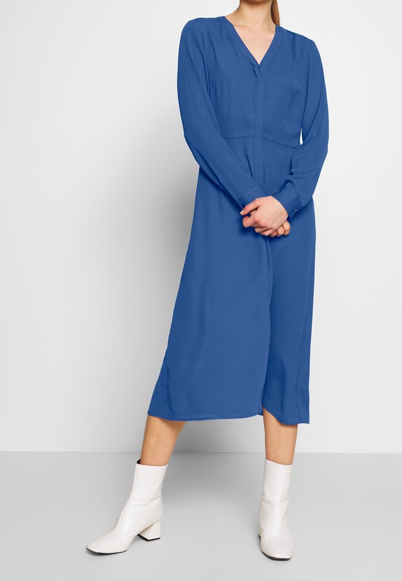 Femme portant une robe midi bleue à manches longues et des bottines blanches, debout, les mains jointes devant, sur un fond uni.