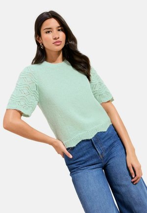 Svetlozelený krátky sveter s textúrovaným pleteným telom a rukávmi s čipkovaným vzorom, v kombinácii s modrými džínsami s vysokým pásom.