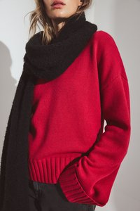 Suéter rojo de punto con dobladillo y puños acanalados, combinado con una bufanda negra texturizada que se drapea alrededor del cuello, acentuando el look en capas.