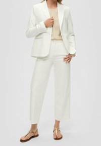 Blazer blanc avec un pantalon large assorti, superposé à un haut beige. Le tissu semble lisse avec un design simple, complété par des sandales dorées en accent.