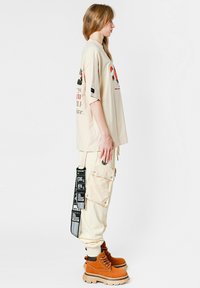 Camisa corta de gran tamaño en beige con estampado gráfico, combinada con pantalones cargo crema con bolsillos y etiquetas negras, calzados con botas marrones de cordones.