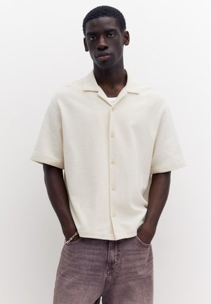 Mand med kort mørkt hår iført en tekstureret lys beige kortærmet skjorte med knapper og falmede lilla jeans, hænderne i lommerne.