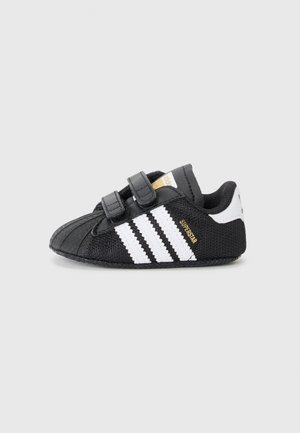 Chaussure bébé Adidas noire avec des bandes blanches et deux bandes velcro, étiquetée "Superstar" en or sur le côté.