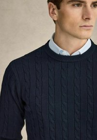 Meripaita tummansinisestä langasta, jossa on pyöreä kaula-aukko ja ribbaus. Paita on puettu vaaleansinisen kauluspaidan päälle, mikä korostaa teksturoitua neulekuviota.