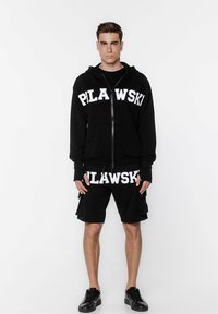 Czarna bluza z kapturem na zamek z napisem "PILAWSKI" białymi literami na piersi, w zestawieniu z pasującymi czarnymi szortami cargo i czarnymi trampkami.