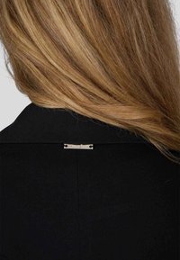 Blazer noir au tissu lisse, avec une étiquette logo argentée à l'arrière près du col, et un design de col classique.