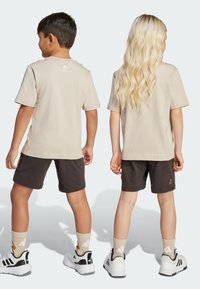 Dos niños de espaldas, vestidos con camisetas beige y pantalones cortos oscuros. Zapatillas blancas y negras; calcetines con un logo. El cabello es claro y castaño.