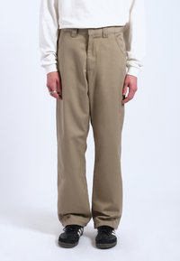 Beige broek van gladde stof met een relaxte pasvorm, voorzien van steekzakken, riemlussen en elastische boorden. gedragen met zwarte sneakers.