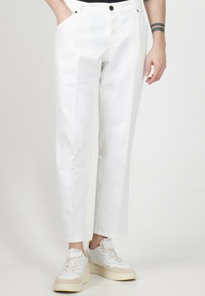 Pantaloni bianchi in cotone con vestibilità dritta, dotati di tasche laterali e chiusura con bottone. Indossati con sneakers di colore chiaro.